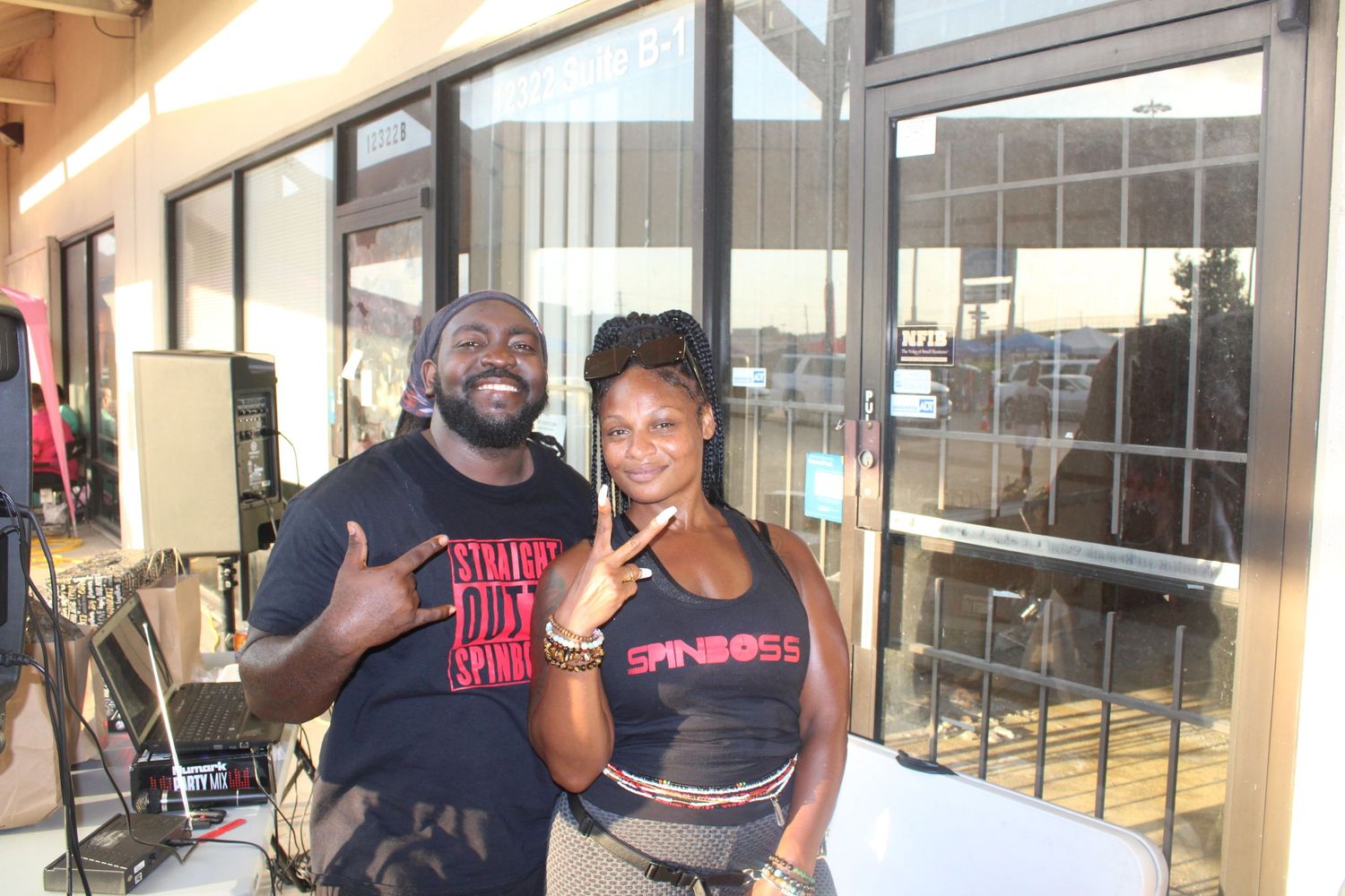 Two people in black t-shirts posing together outside a storefront with smiles and friendly gestures.