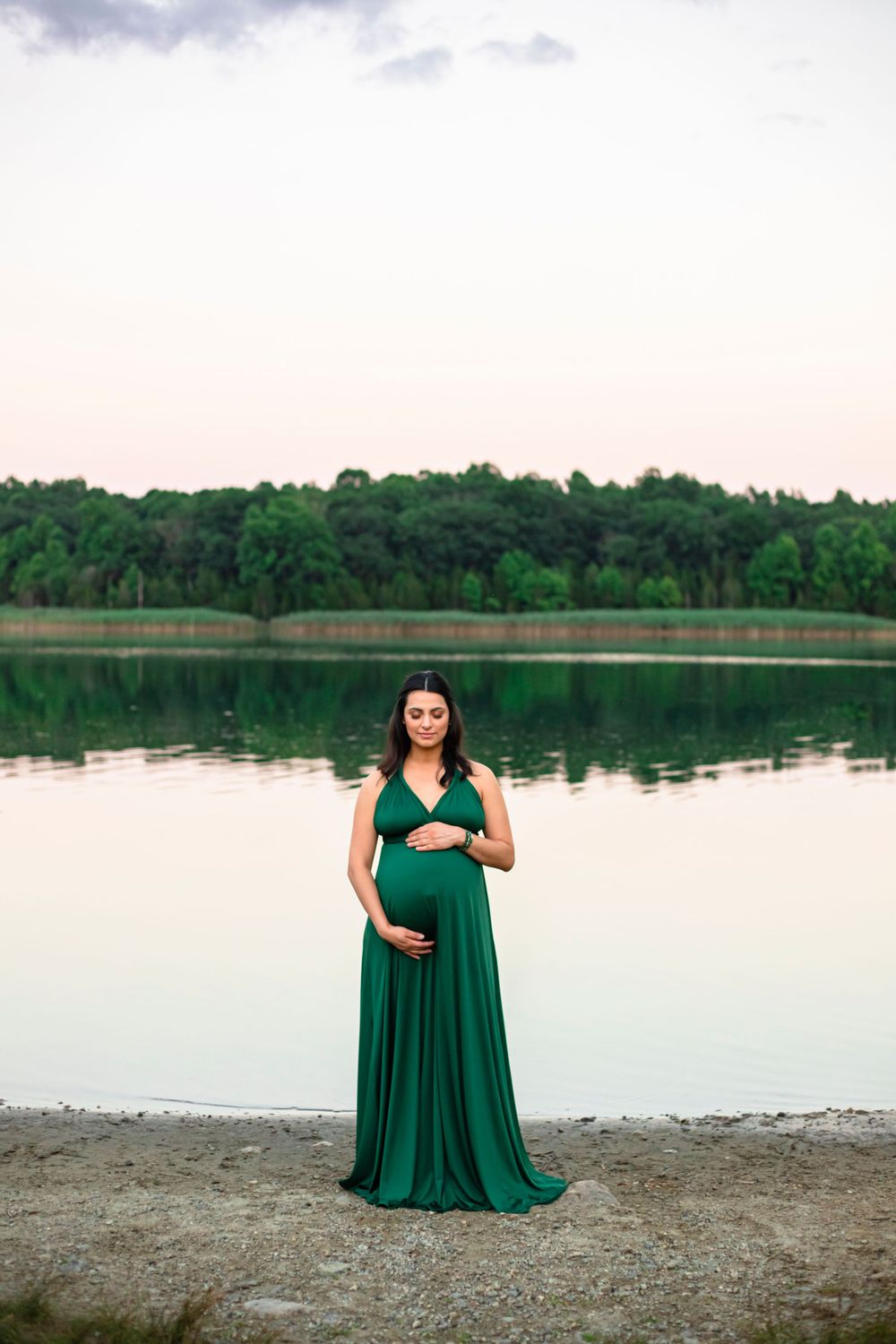Maternity photo of woman in emerald green dress standing by serene lake at sunset.