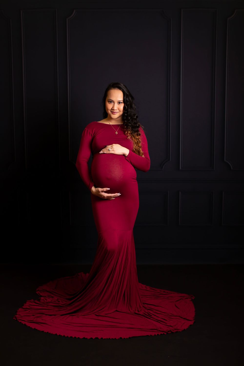 A dramatic maternity portrait of an expectant mother in a flowing burgundy gown against a dark background.