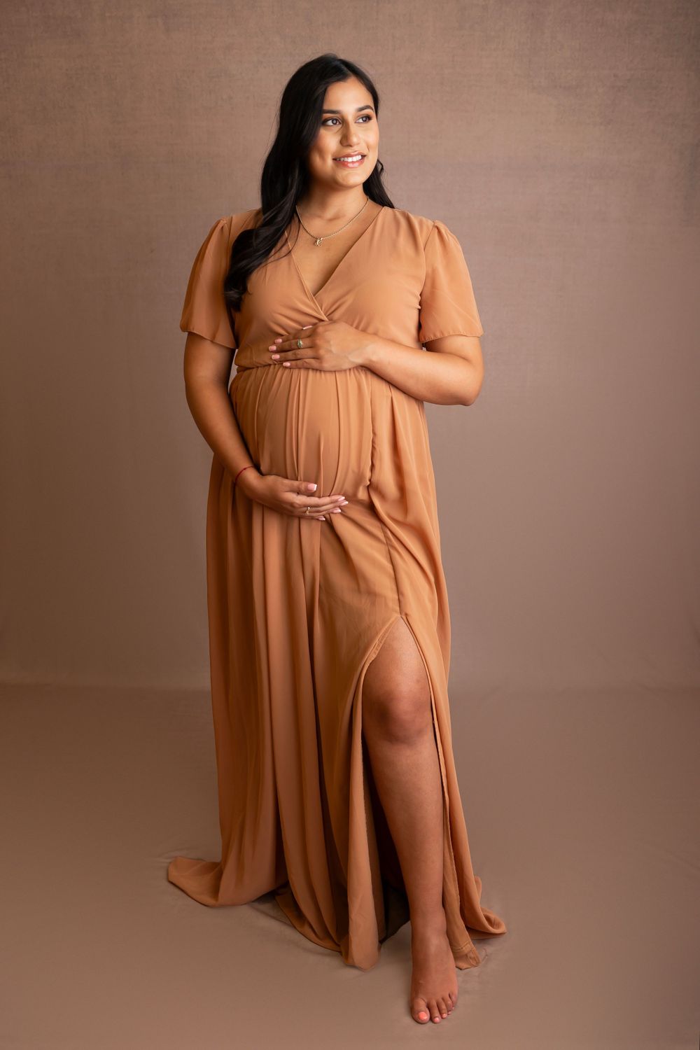 Elegant maternity portrait in flowing terracotta dress against neutral backdrop.