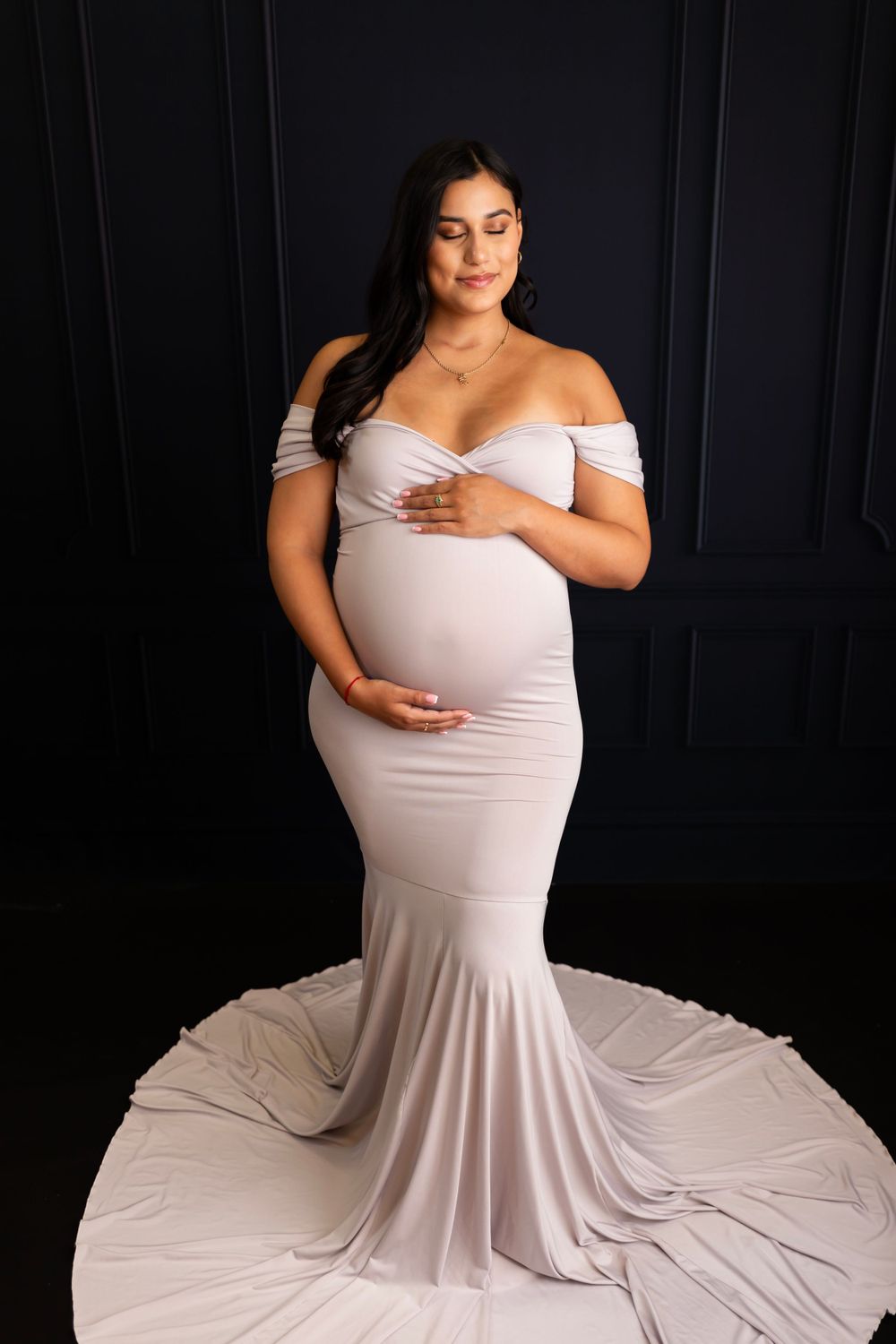 Dramatic maternity photo in off-shoulder white mermaid gown against dark background.