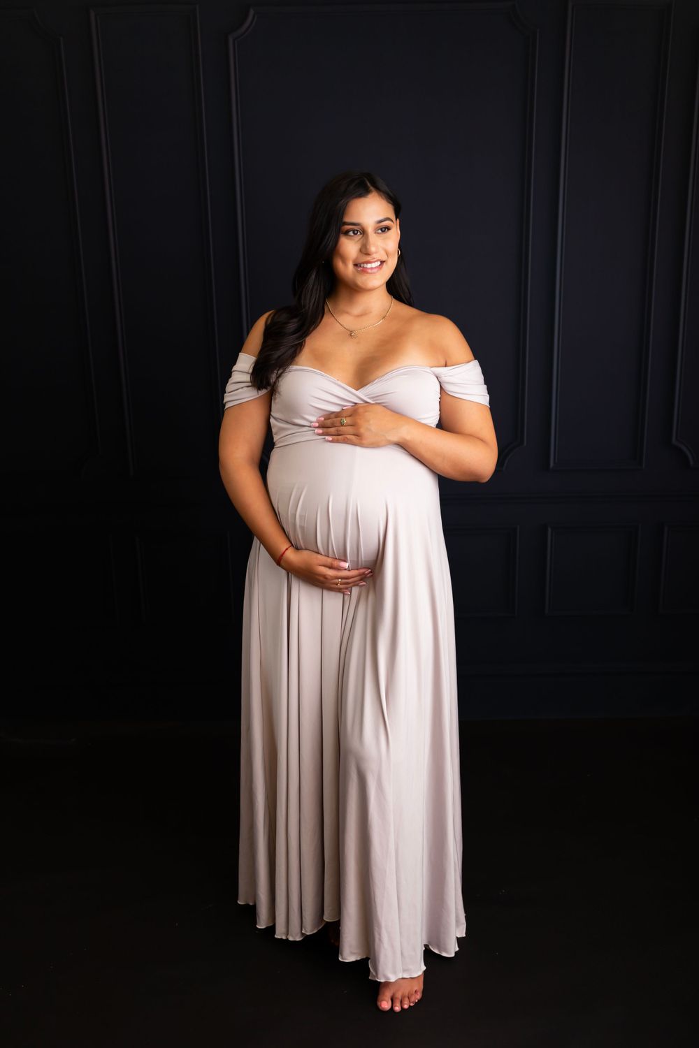 Elegant maternity portrait in floor-length white off-shoulder dress against dark wall.