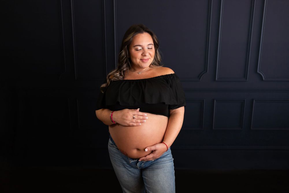 Casual maternity portrait in black top and jeans against navy wall.