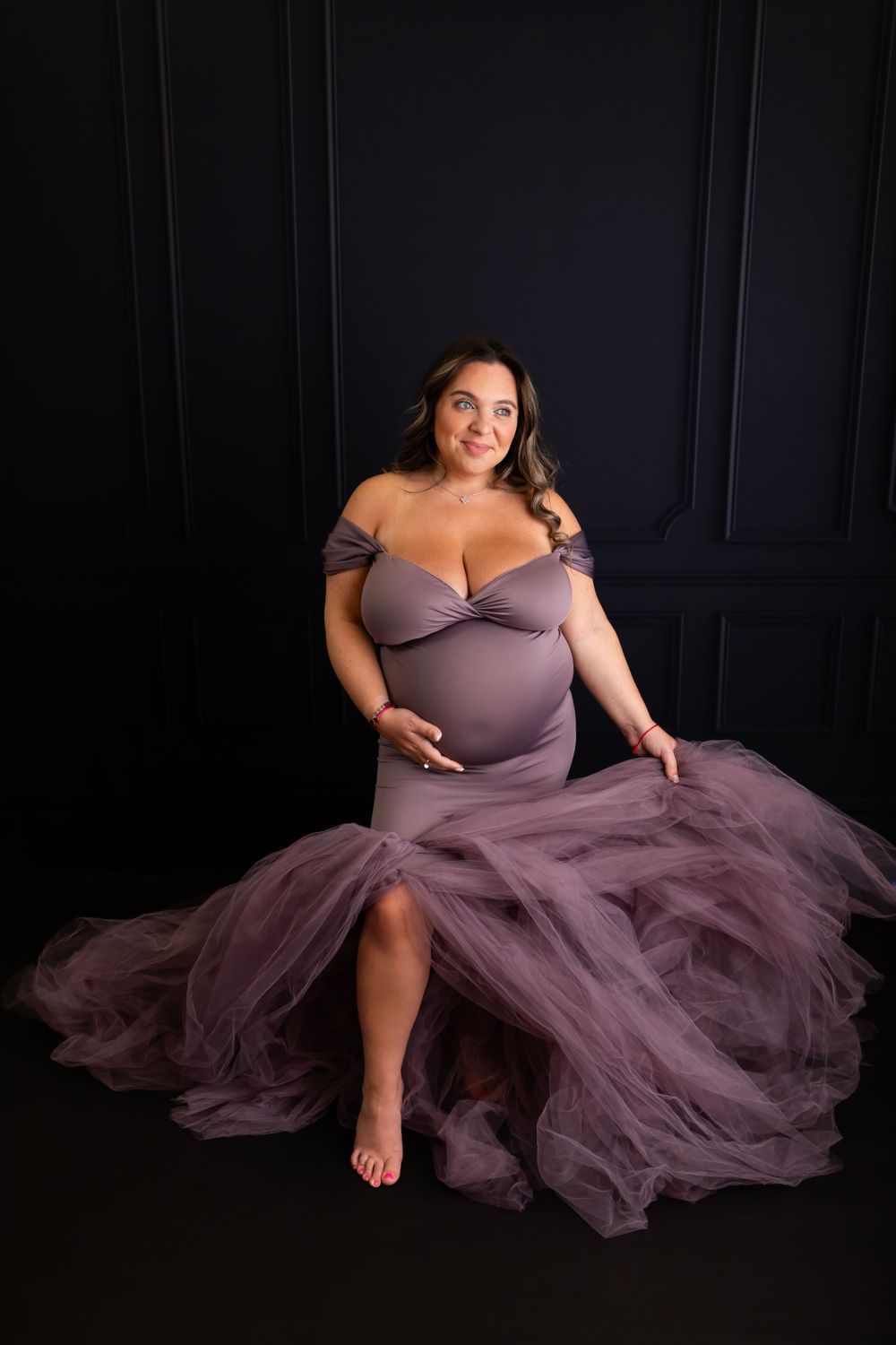 Maternity portrait in flowing purple tulle gown against dark backdrop.