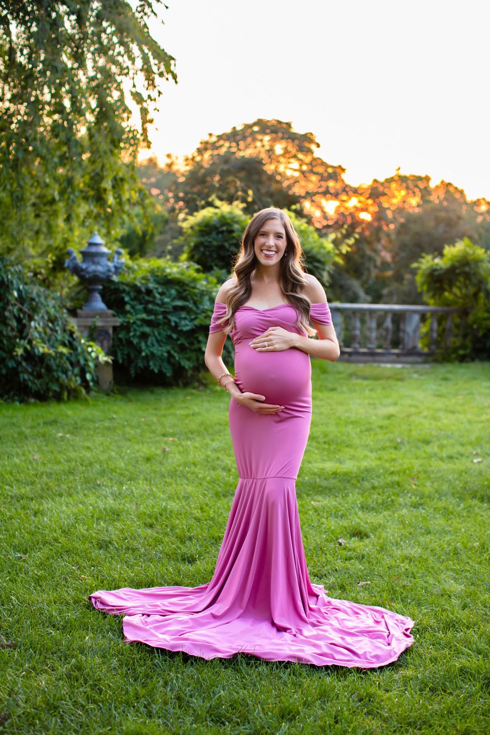 Stunning maternity photo of woman in pink flowing dress against garden backdrop.