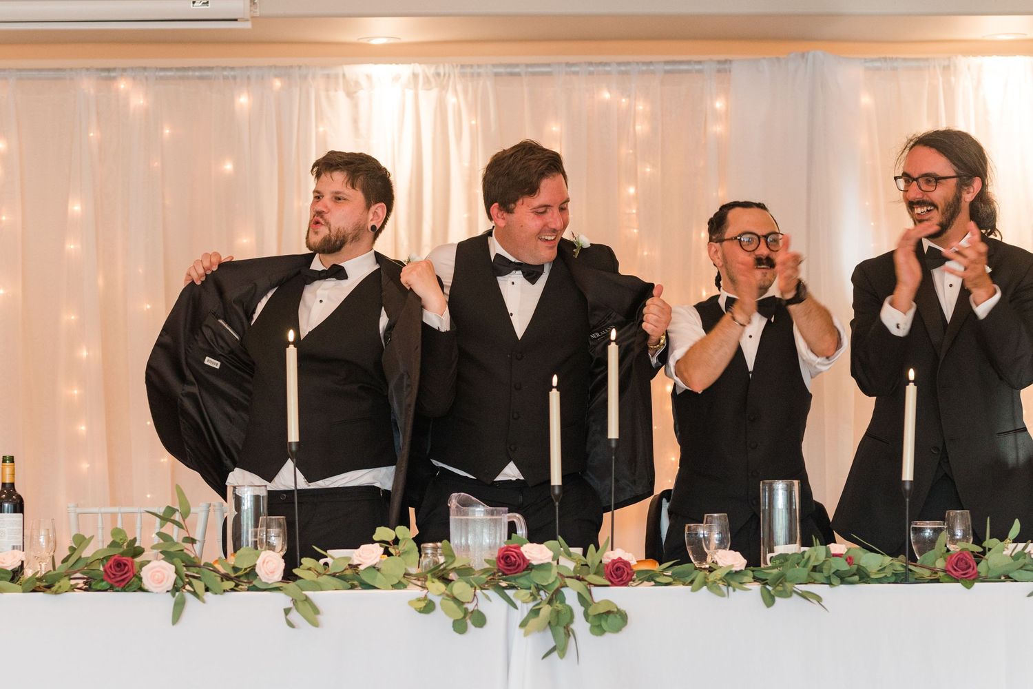 Wedding party members in formal attire seated at decorated head table during reception celebration.