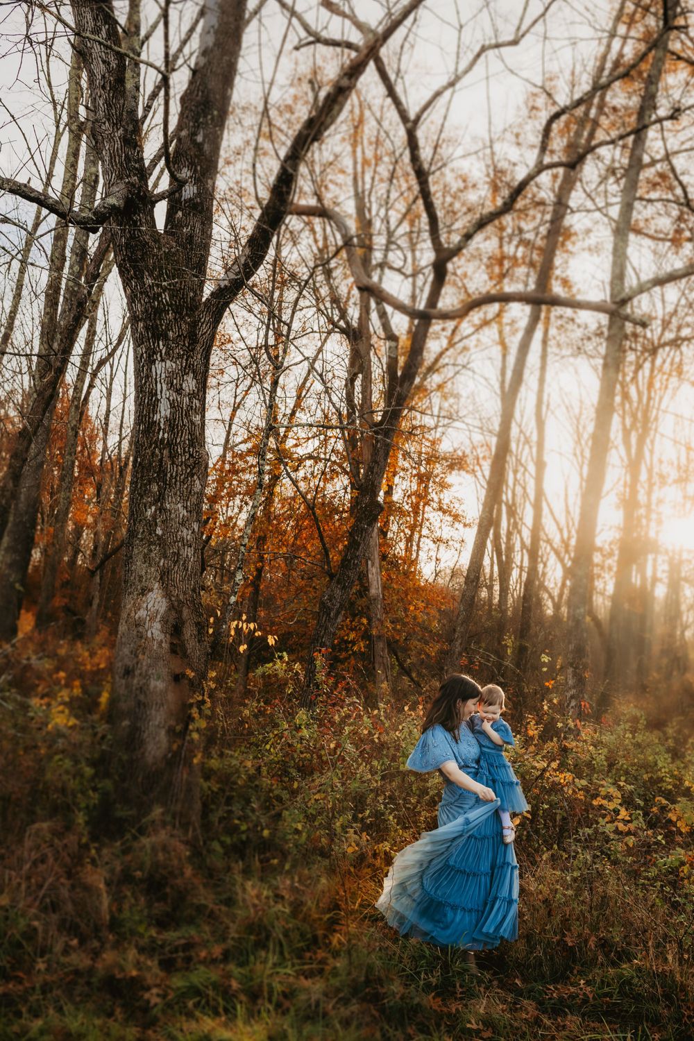 A figure in a flowing blue dress twirls through an autumn forest at sunset.