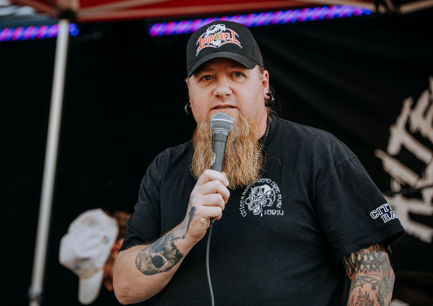 A bearded performer in a black t-shirt and baseball cap holds a microphone during an outdoor performance.