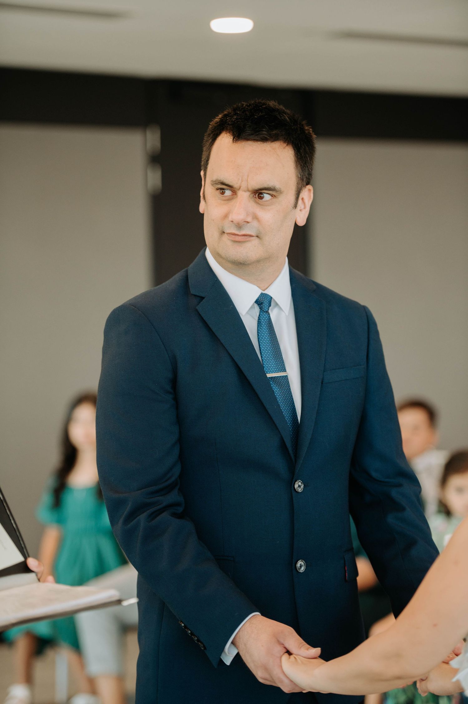 Groom in a navy suit and blue tie stands during wedding ceremony.