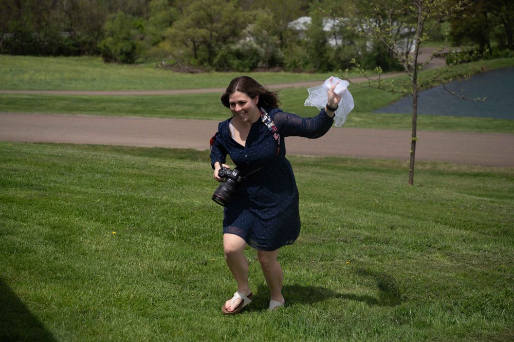 Someone in a blue dress dances joyfully outdoors on grass with a lake behind them.