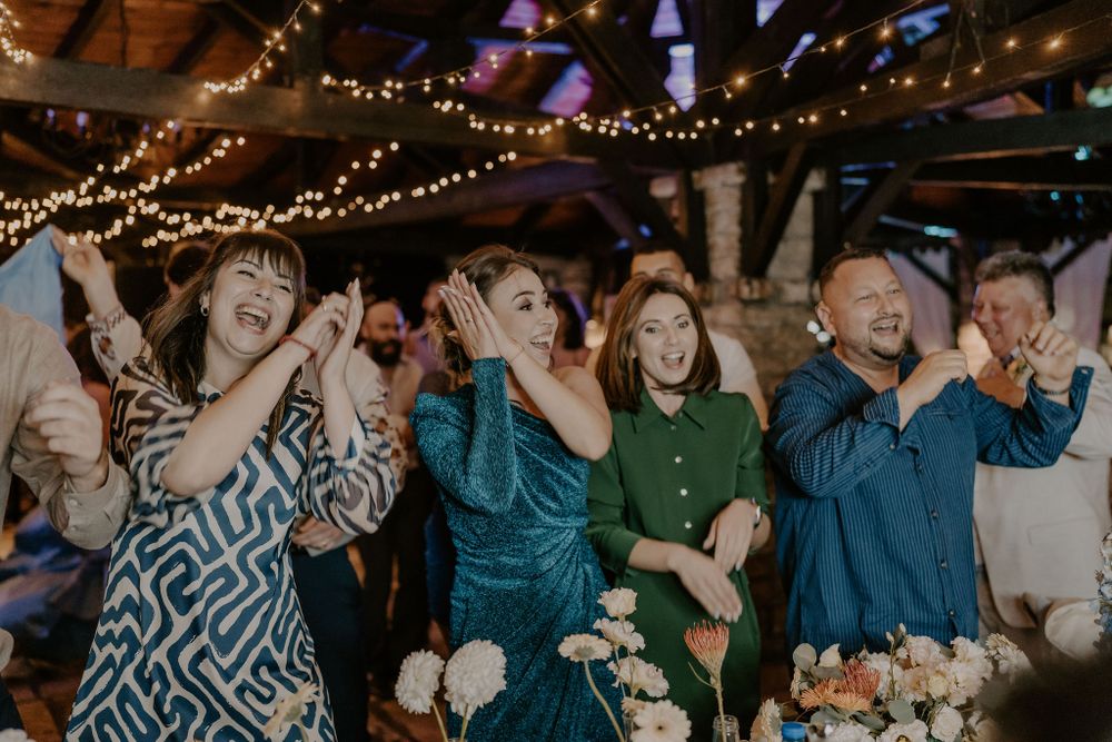 Wedding guests dancing and celebrating under rustic string lights at an indoor reception venue.