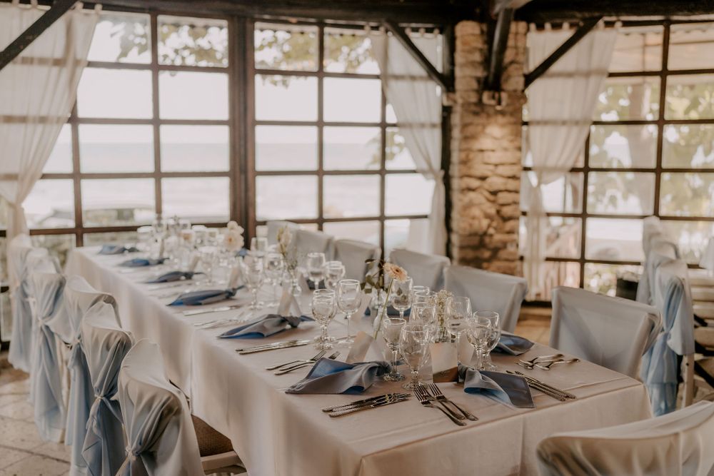 Long dining table with white tablecloth and blue napkins set for formal occasion in rustic venue.