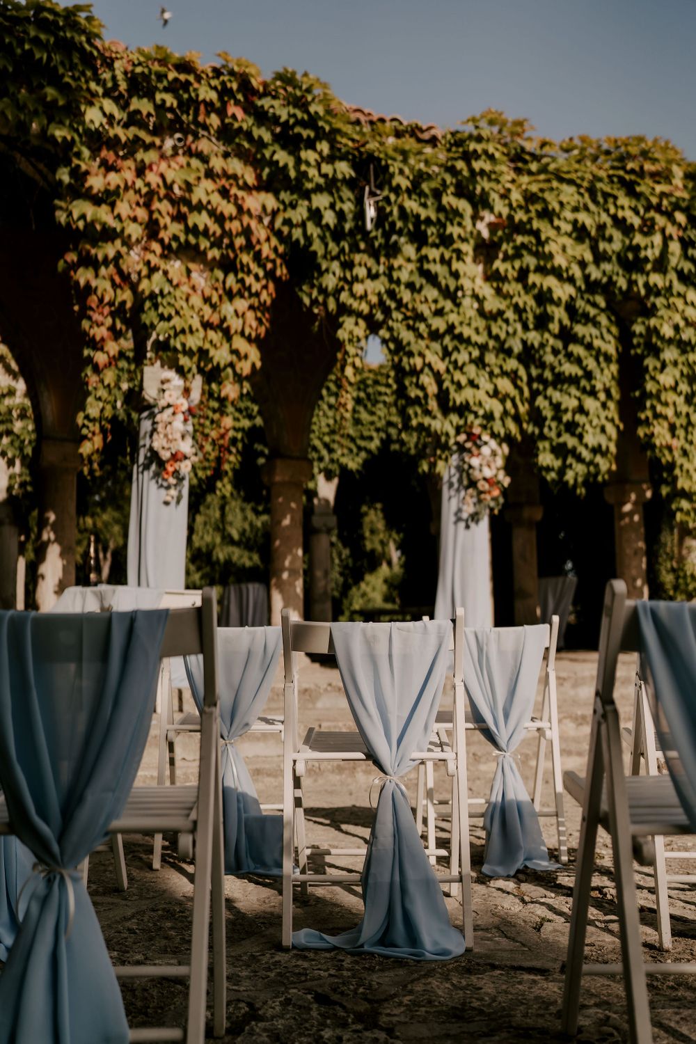 White ceremony chairs with dusty blue fabric ties arranged in rows under trees with fall colors.