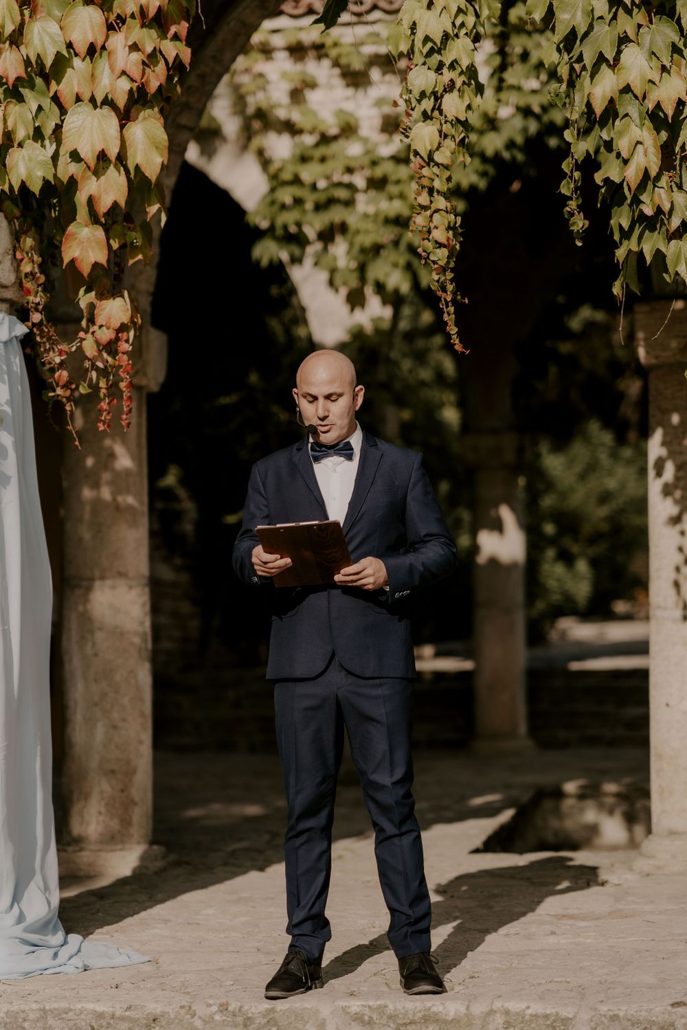 Wedding officiant in dark suit stands under ivy-covered archway during ceremony.
