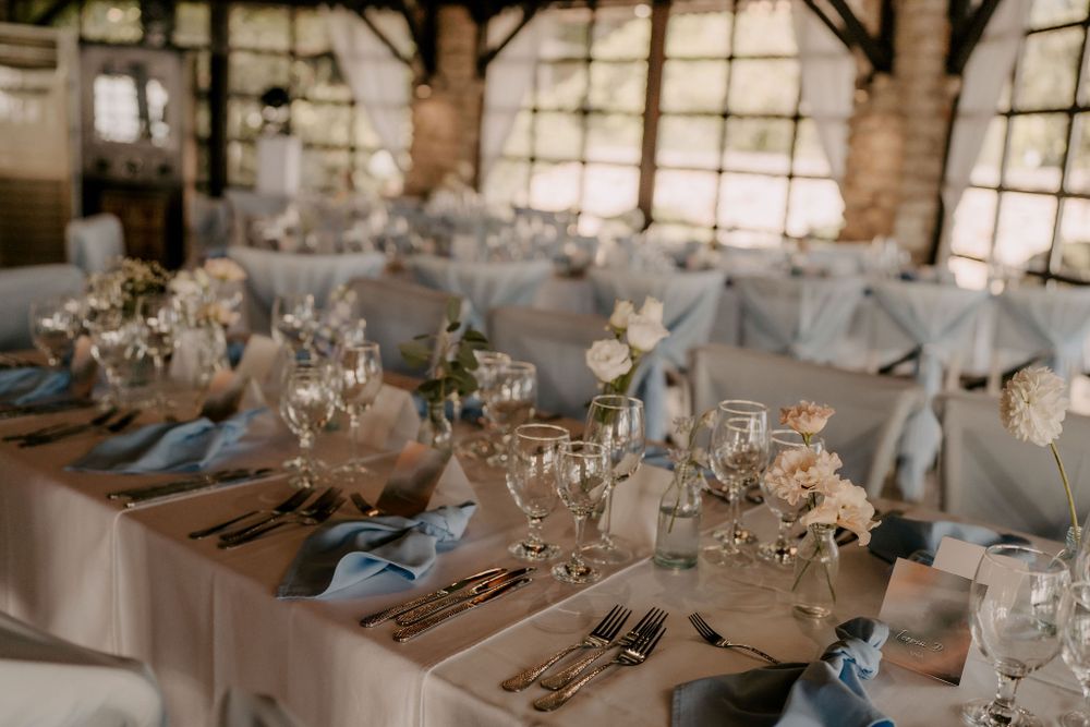 Elegant wedding reception tables with blue accents and floral centerpieces in sunlit venue.