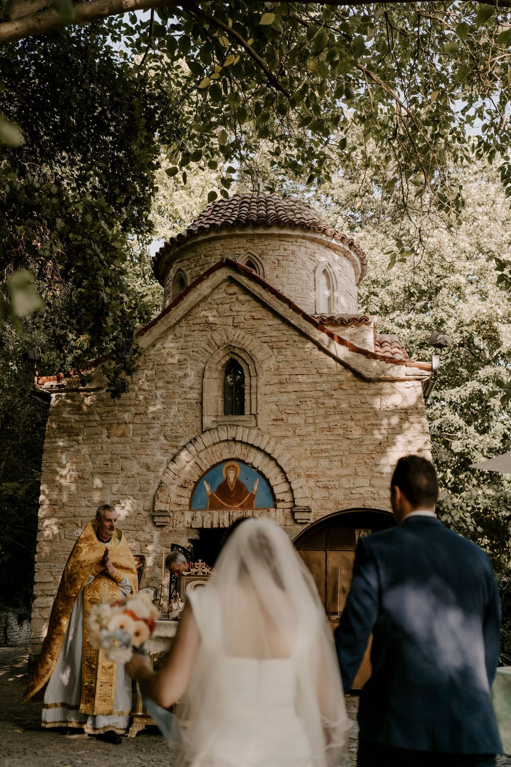 A wedding ceremony procession outside a charming stone chapel with arched doorways.