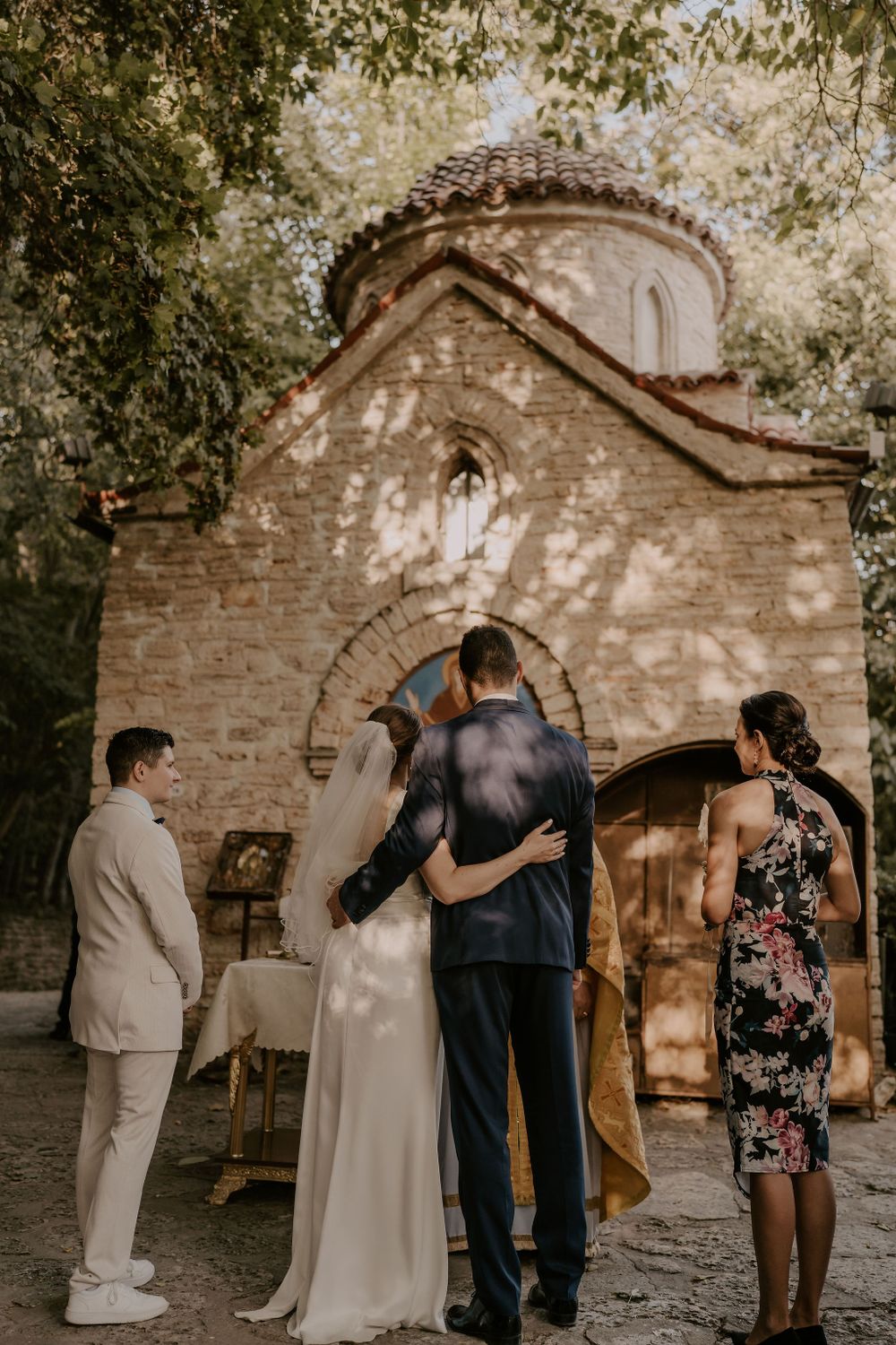 A wedding party gathers outside a rustic stone chapel during a summer ceremony.