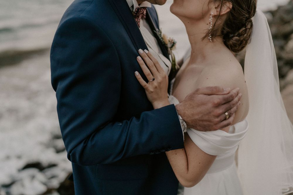 A romantic wedding couple embraces on a rocky beach shoreline during an intimate photo session.