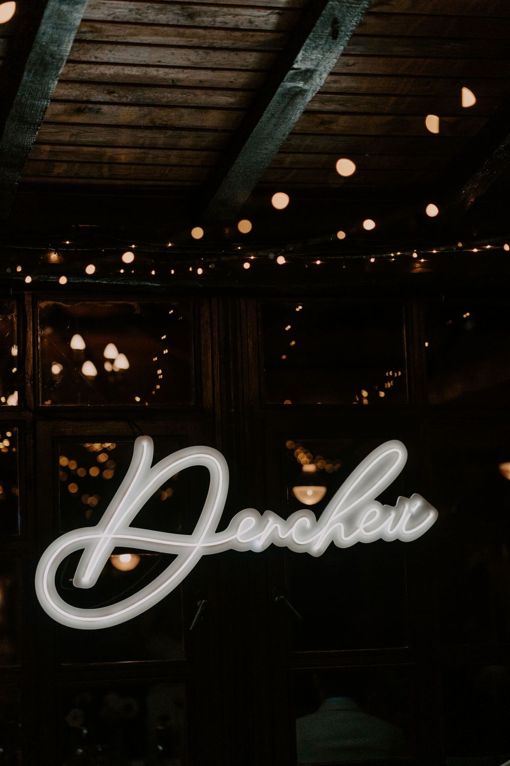 Illuminated cursive neon sign against dark wooden beams with string lights.