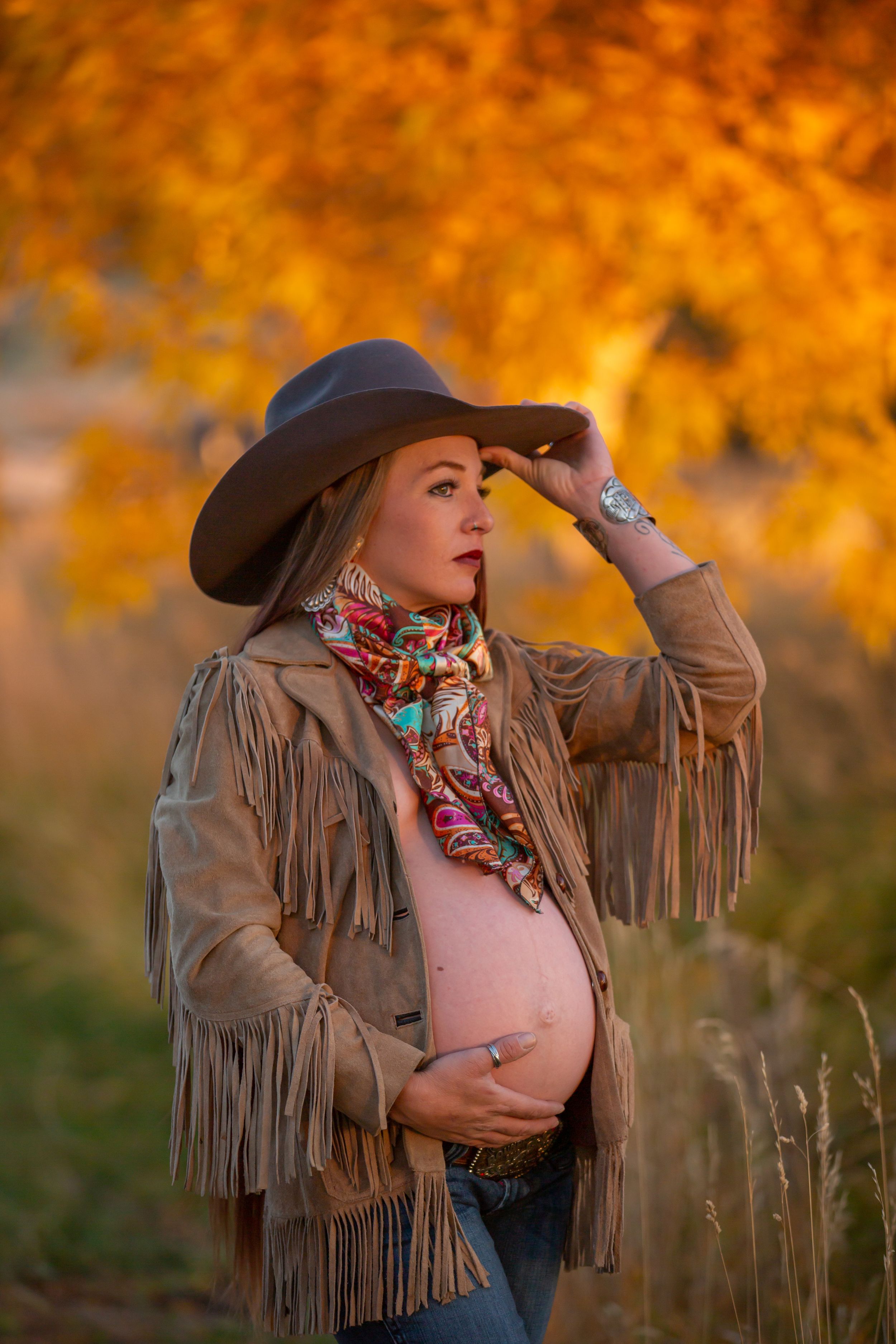 Pregnant woman in fringe jacket and cowboy hat poses in golden autumn field.