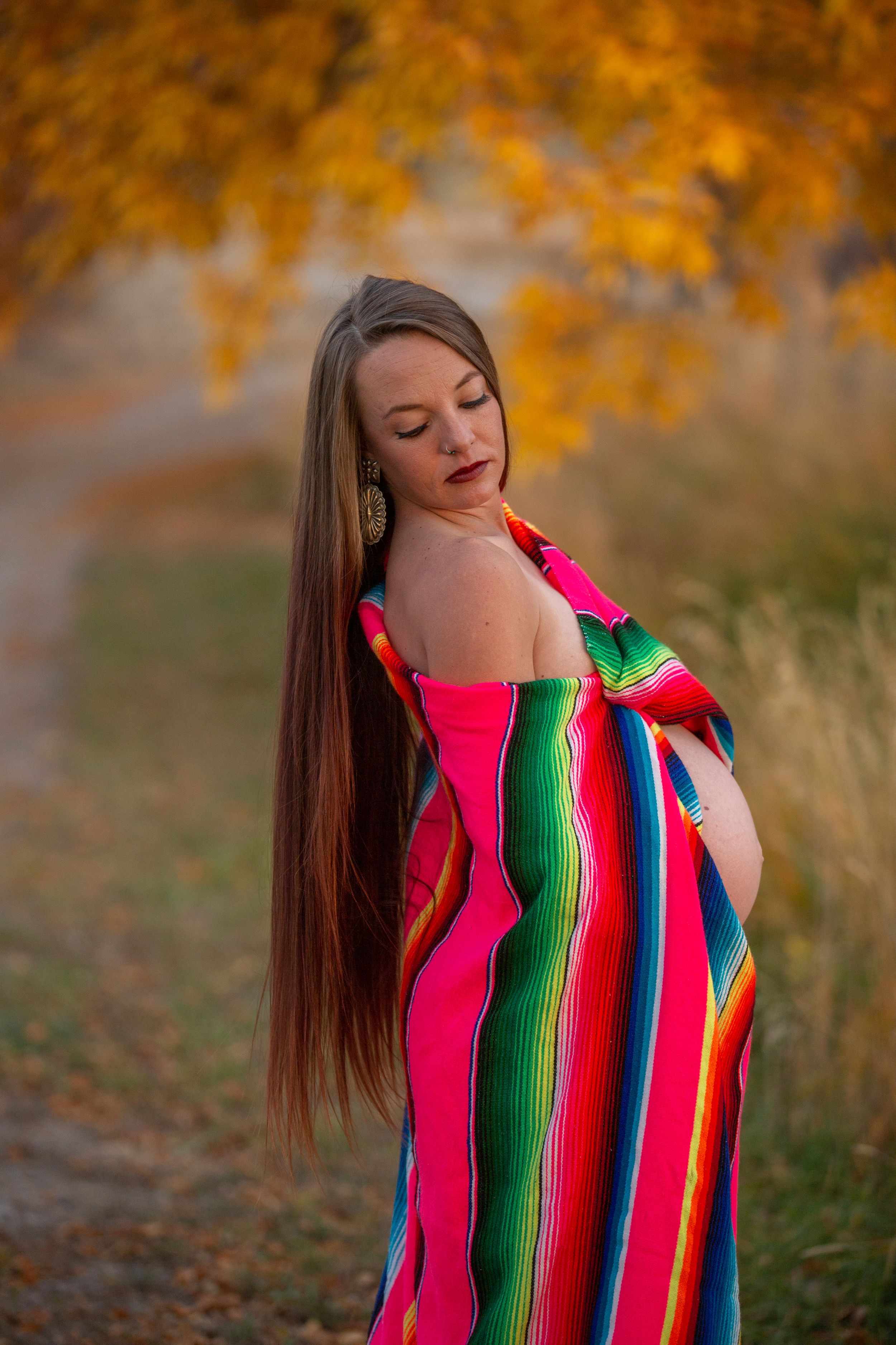 Pregnant woman in colorful striped serape wrap poses on rural path in fall.