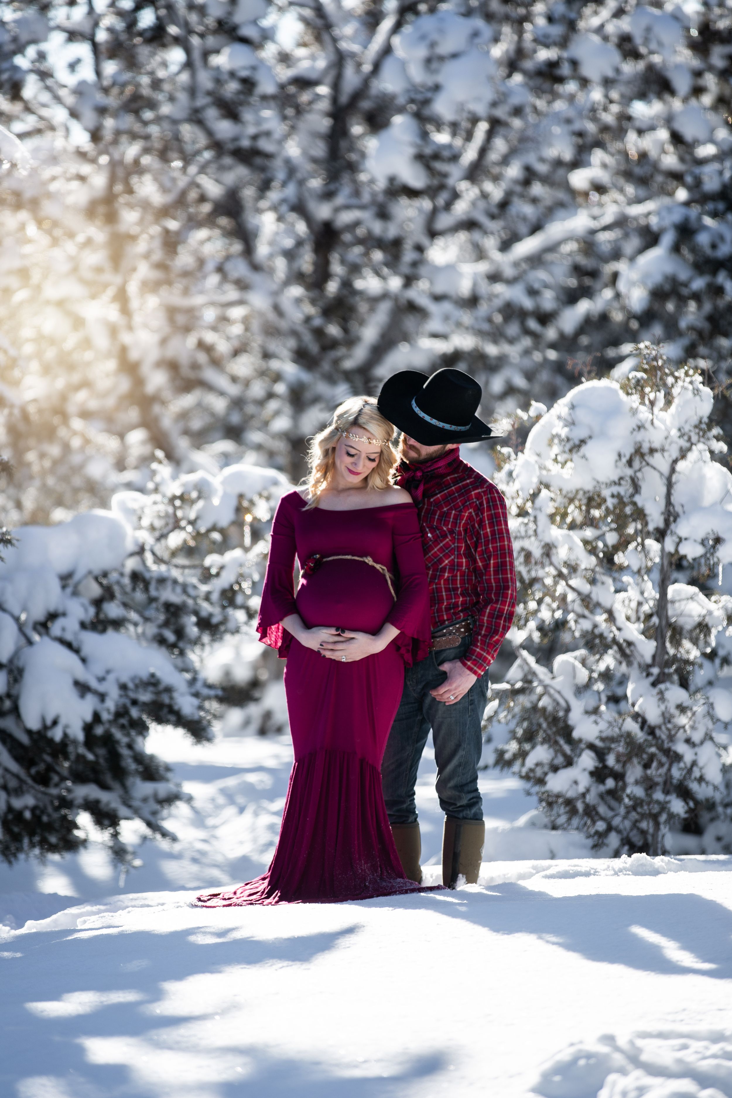 Couple in snowy winter forest, woman in burgundy maternity gown with cowboy partner.