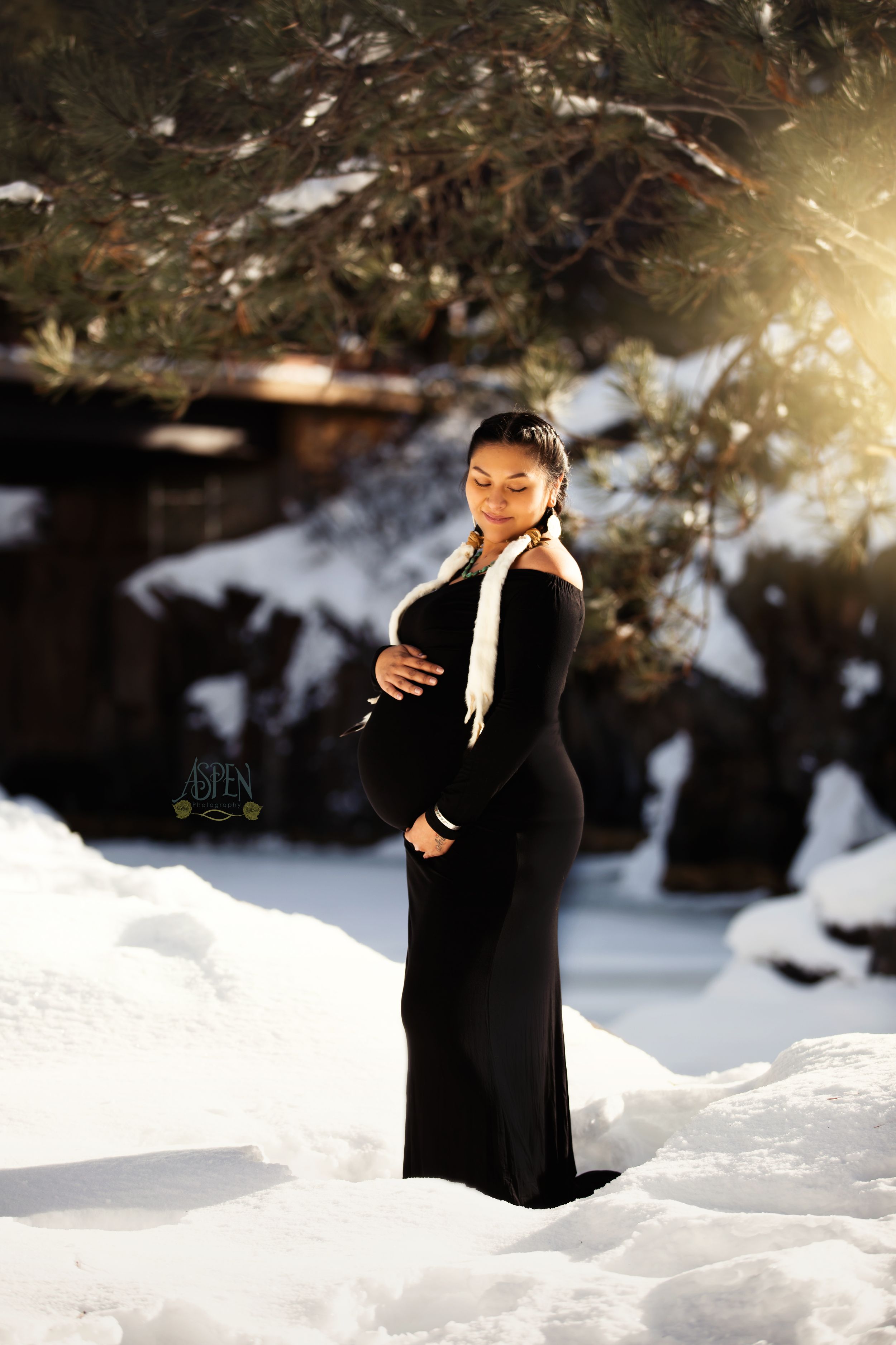 Pregnant woman in black gown holding baby bump in snowy winter outdoor maternity photo session.