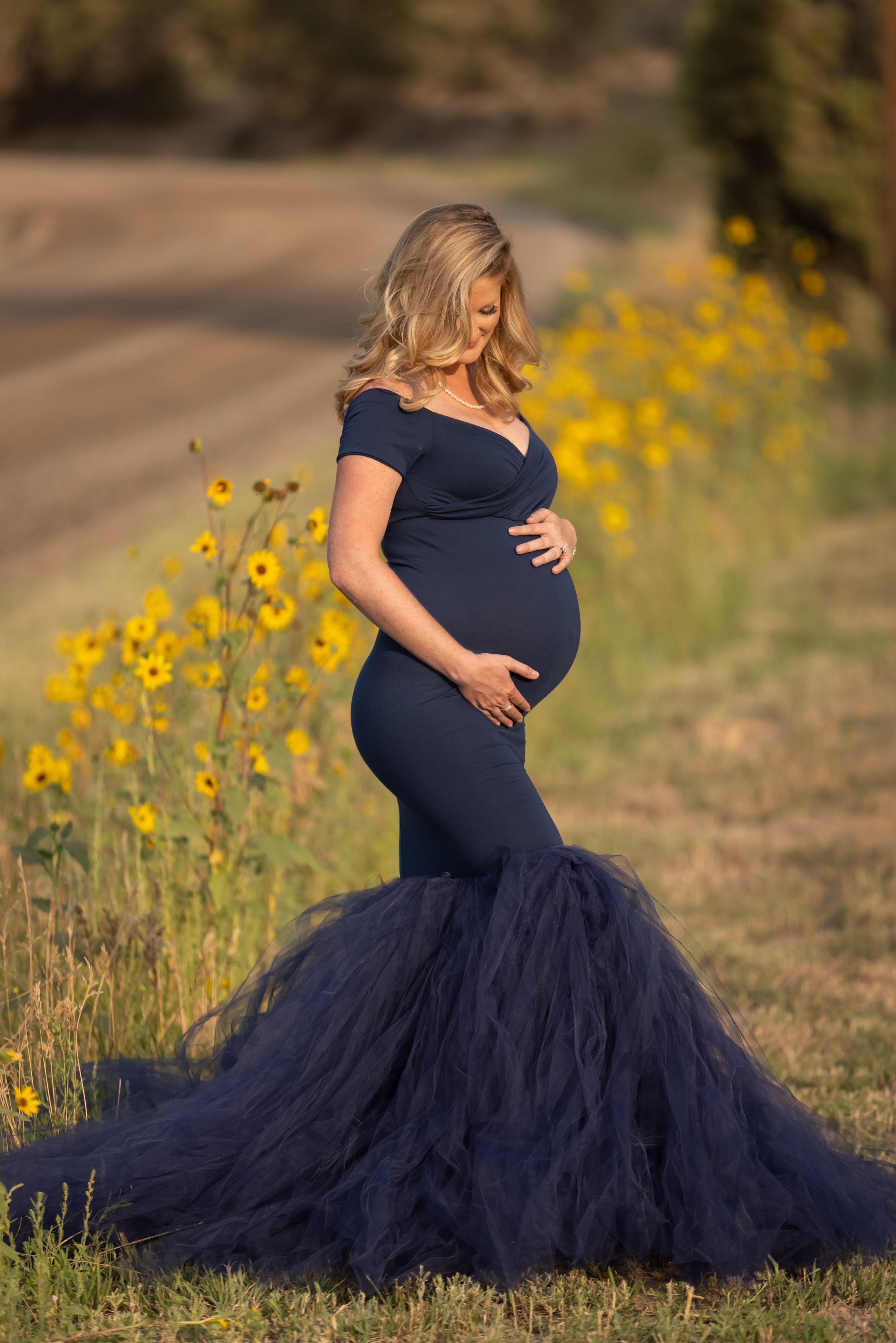 Blonde pregnant woman in navy blue mermaid tulle gown posing beside sunflowers in golden hour light.