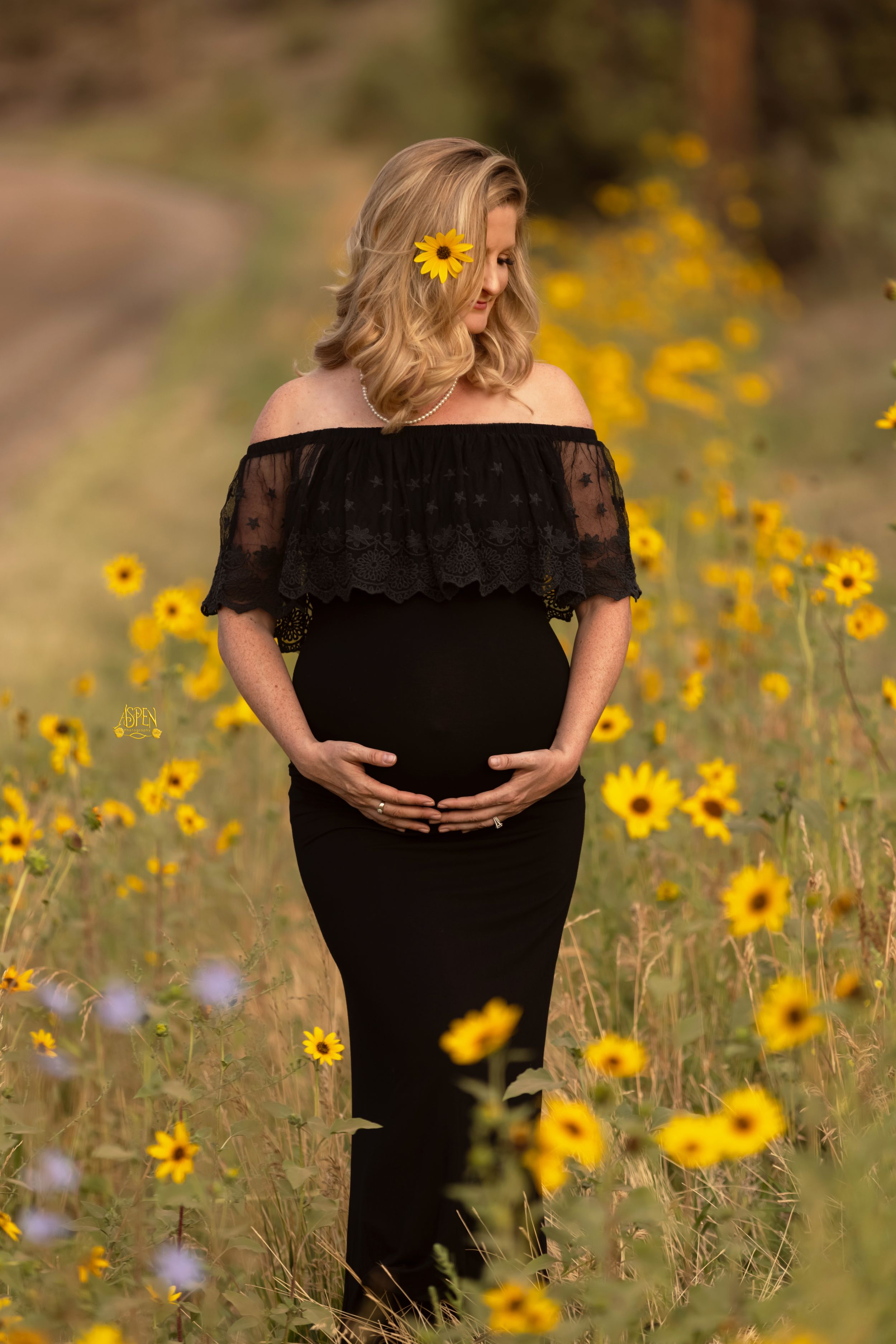 Pregnant woman in black off-shoulder lace dress cradling baby bump in sunflower field maternity shoot.