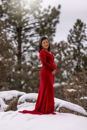 Pregnant woman in elegant red velvet gown standing on snow-covered rocks surrounded by pine trees.