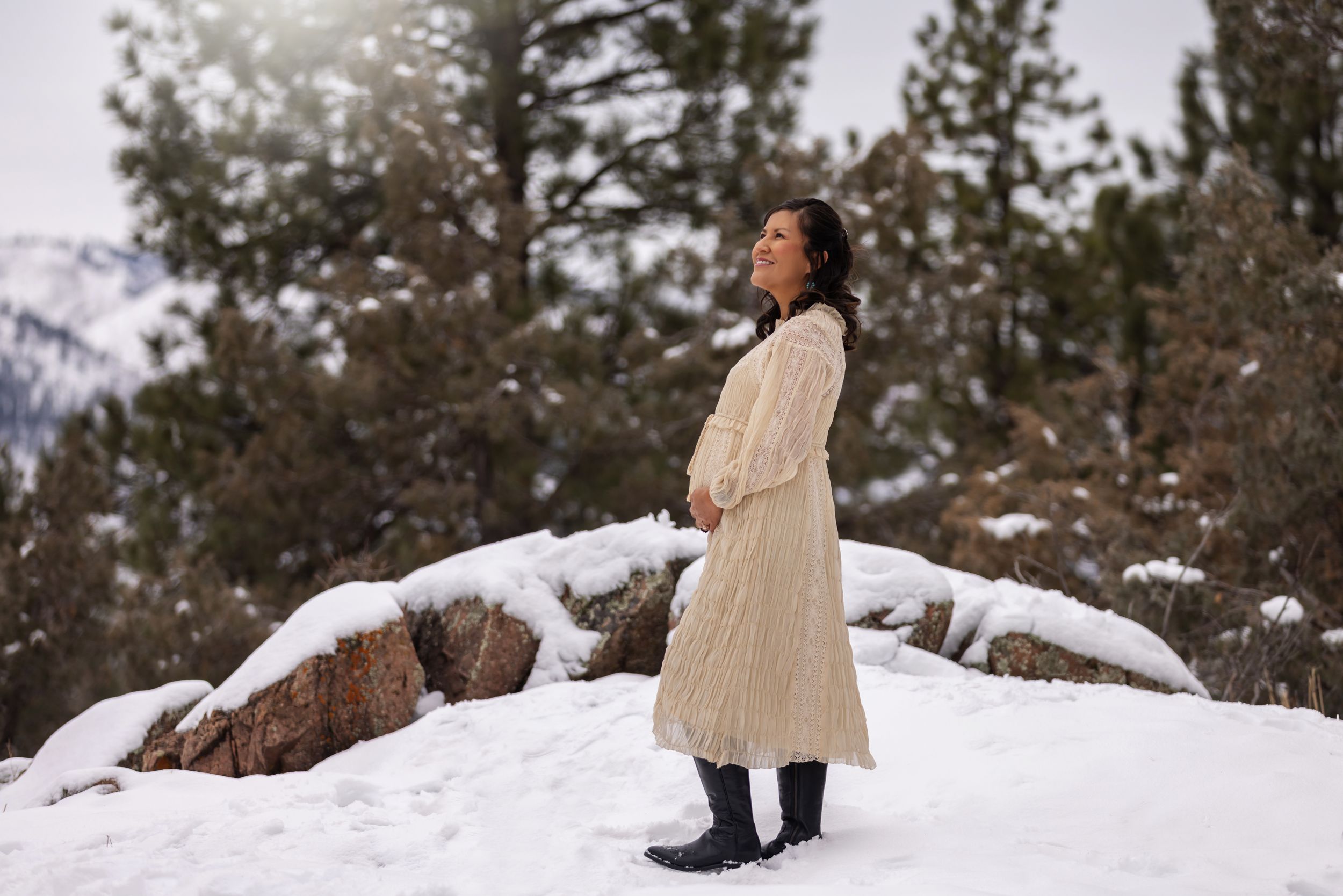 Pregnant woman in cream knit coat standing peacefully in snowy mountain landscape with pine trees.