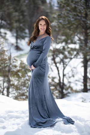 Smiling pregnant woman in gray gown stands in snowy winter forest landscape.