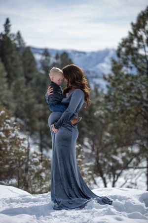 Pregnant woman in gray gown holds toddler in snowy mountain forest setting.