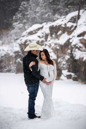 Couple embraces in snowy landscape, man in cowboy hat, woman in white lace gown.