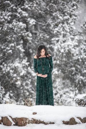 Pregnant woman in emerald gown stands peacefully in heavy snowfall among pines.
