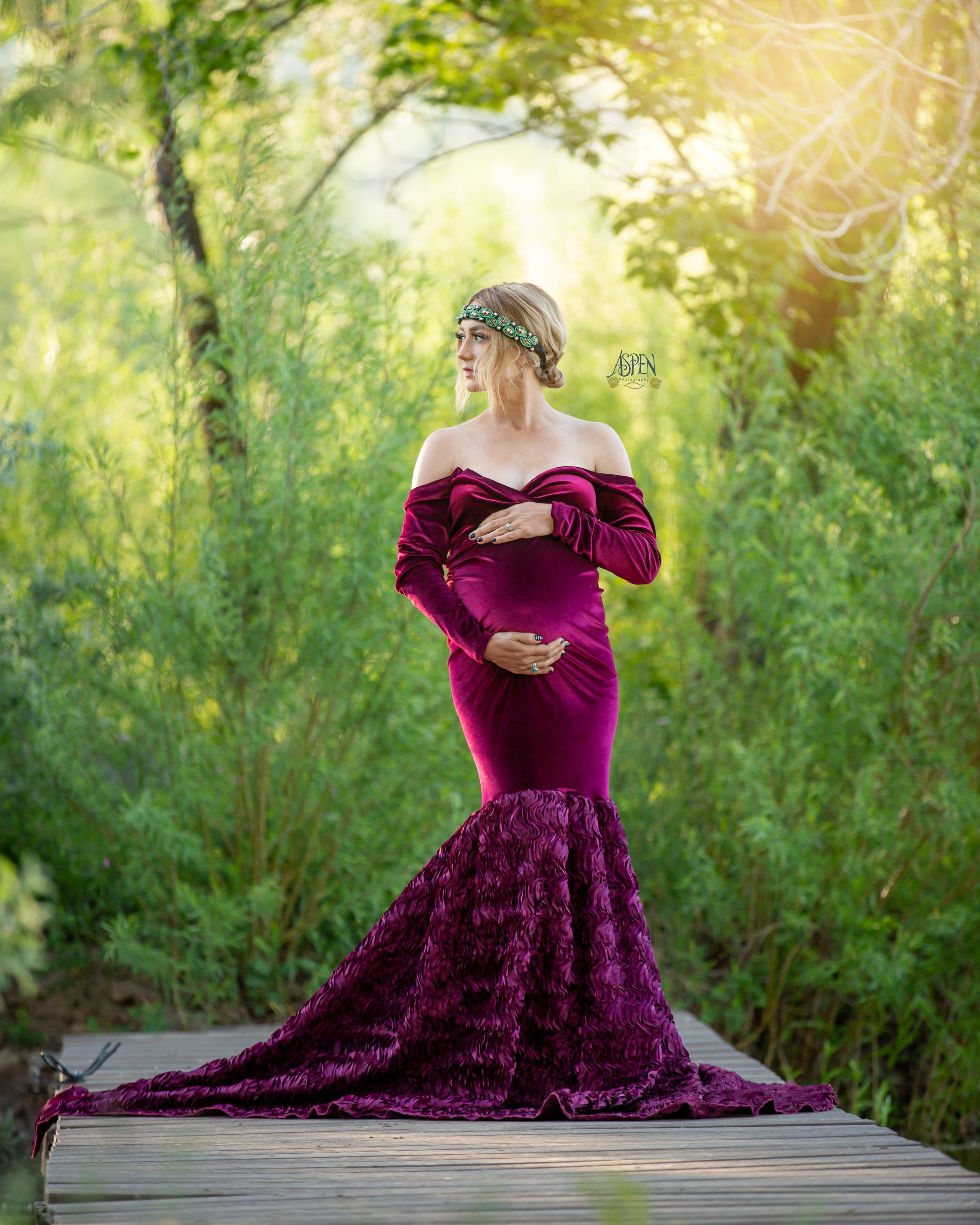 Elegant pregnant woman in purple velvet mermaid gown posing outdoors in lush green garden setting.