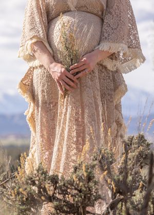 Pregnant woman in cream lace maternity gown holding wildflowers in desert landscape with mountains.