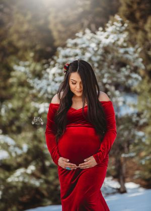 Pregnant woman in red velvet gown poses in sunlit snowy pine forest.