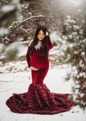 Pregnant woman in burgundy mermaid gown poses in sunlit snowy winter forest.