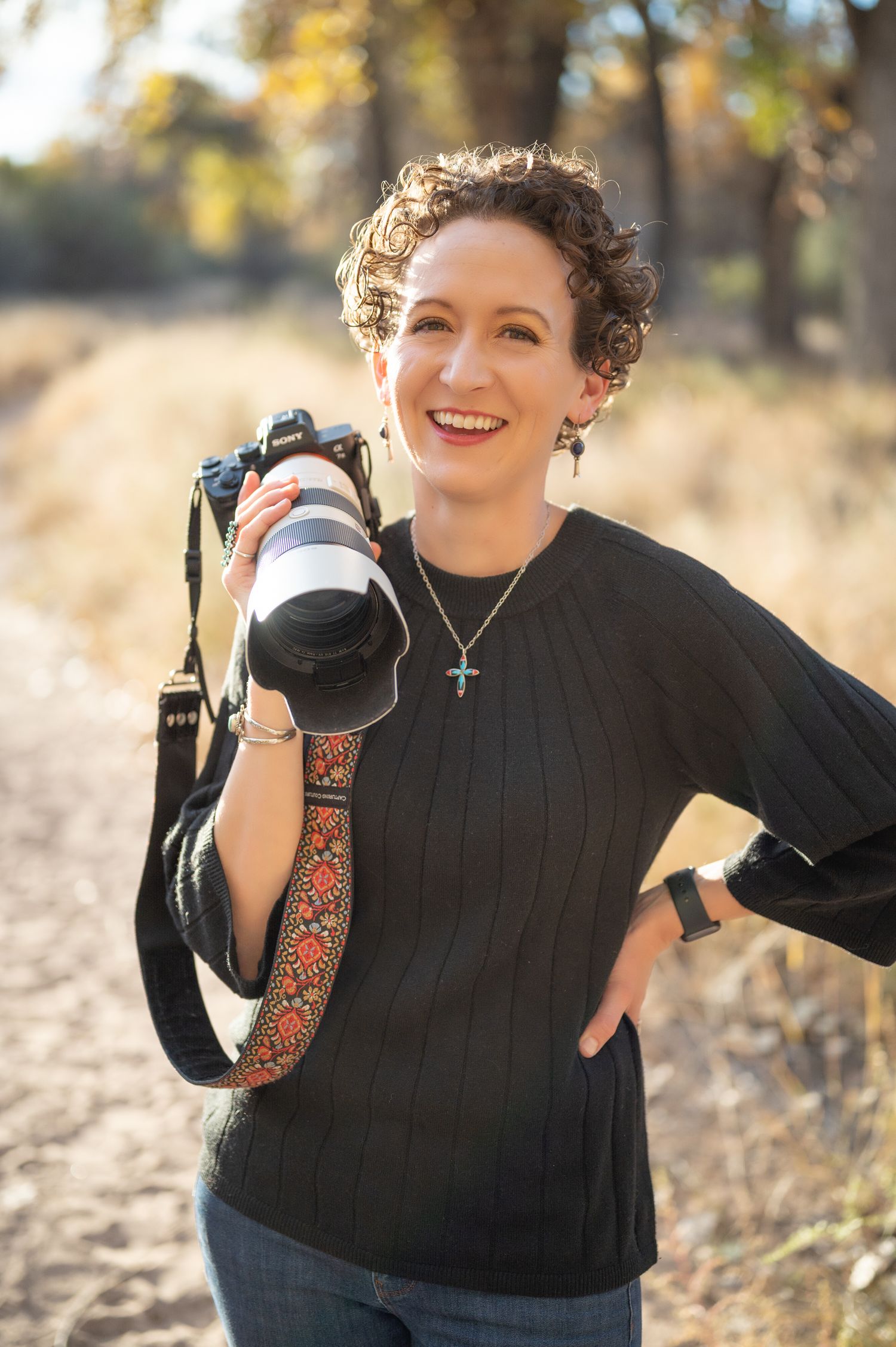 Photographer in black top shows camera while posing outdoors in fall scenery.