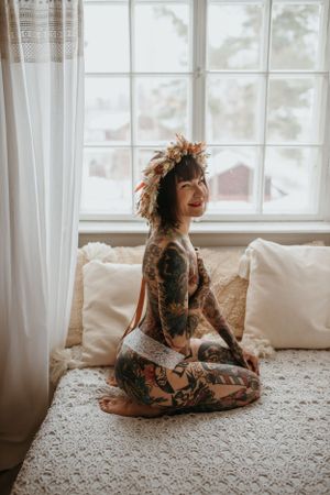 Boheemi boudoir-kuvaus ikkunavalossa, tatuoitu nainen kukkaseppele päässä
