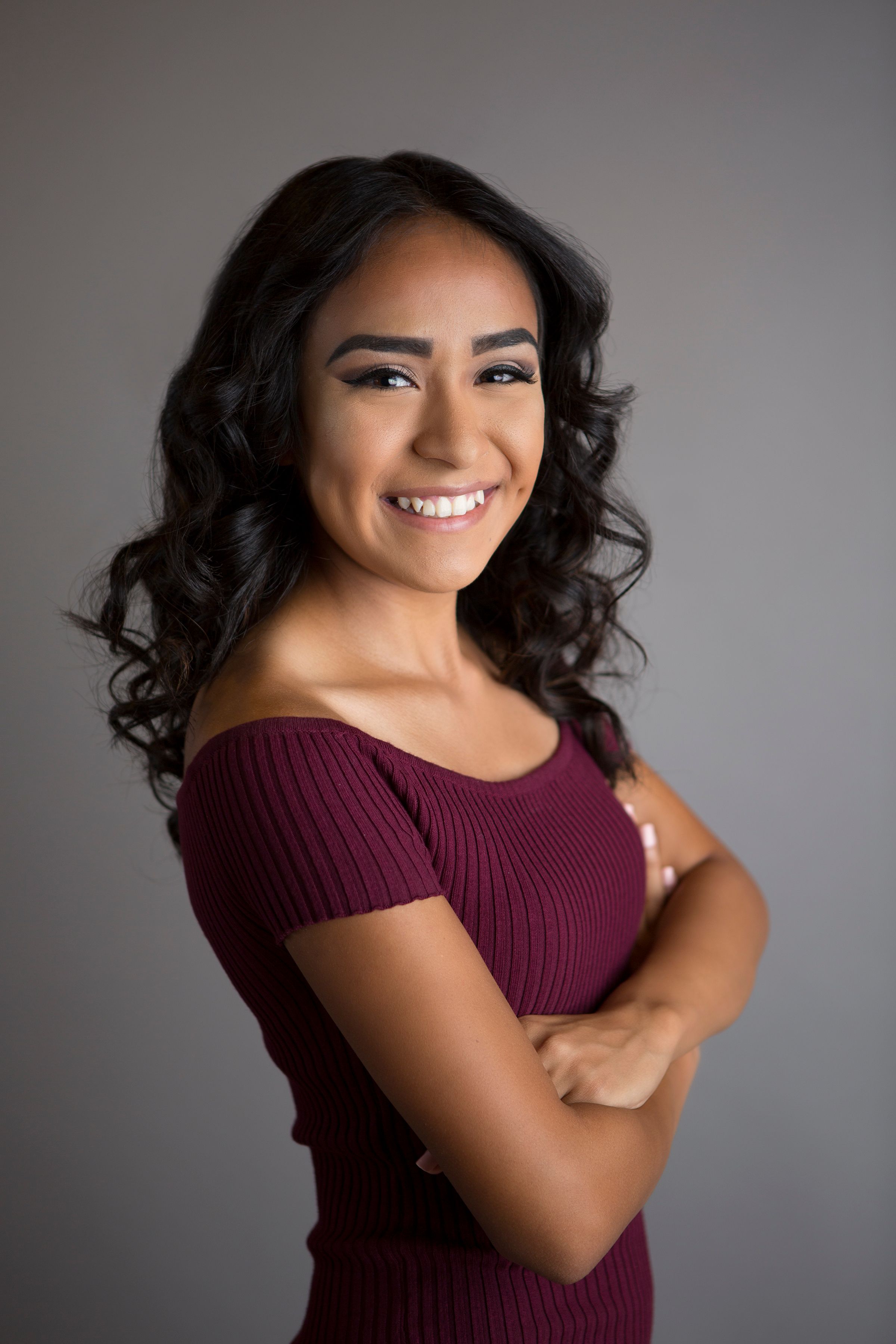 Studio portrait of someone in a burgundy top with curly dark hair against a gray background.