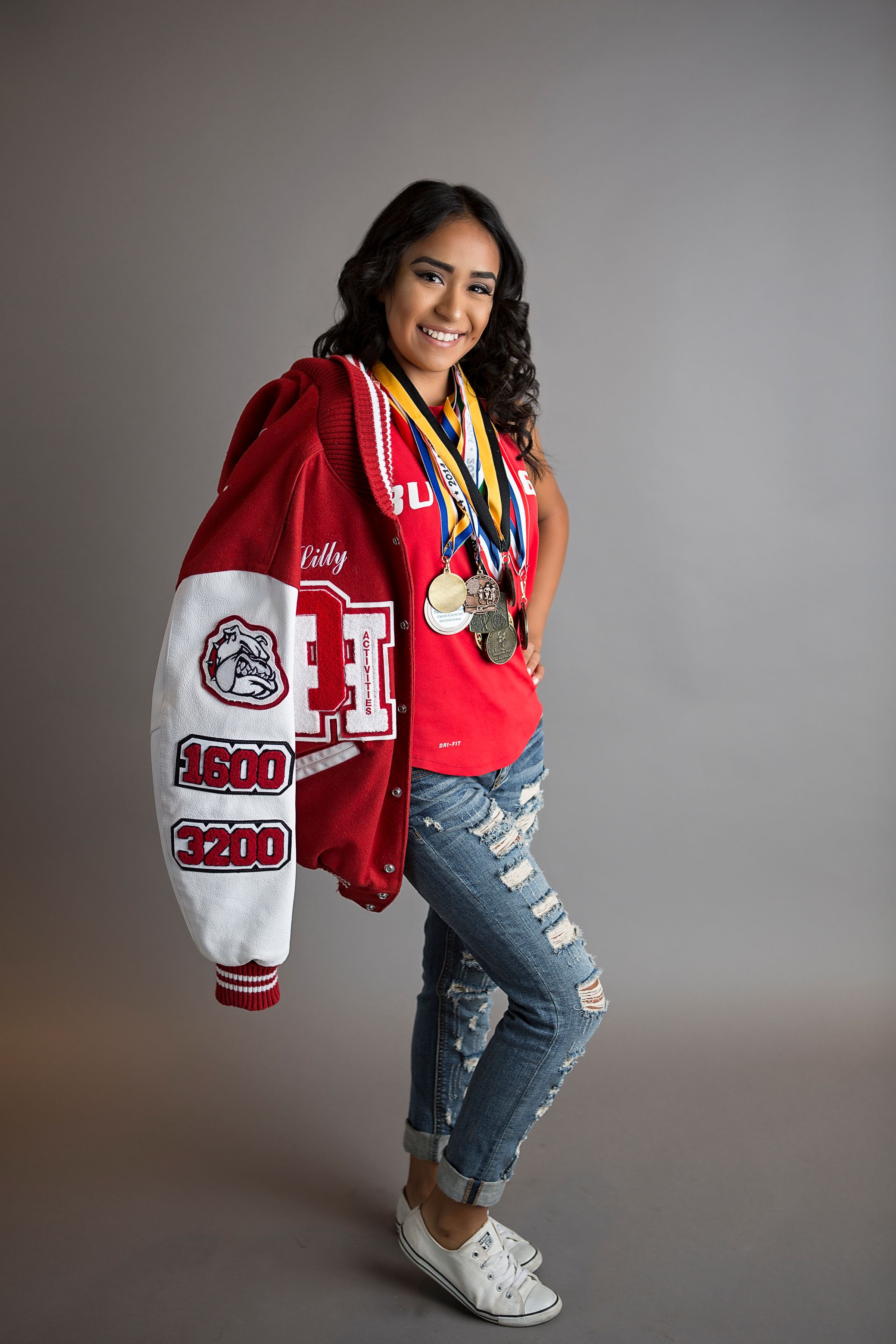 Student wearing varsity jacket and ripped jeans poses with athletic gear against gray backdrop.