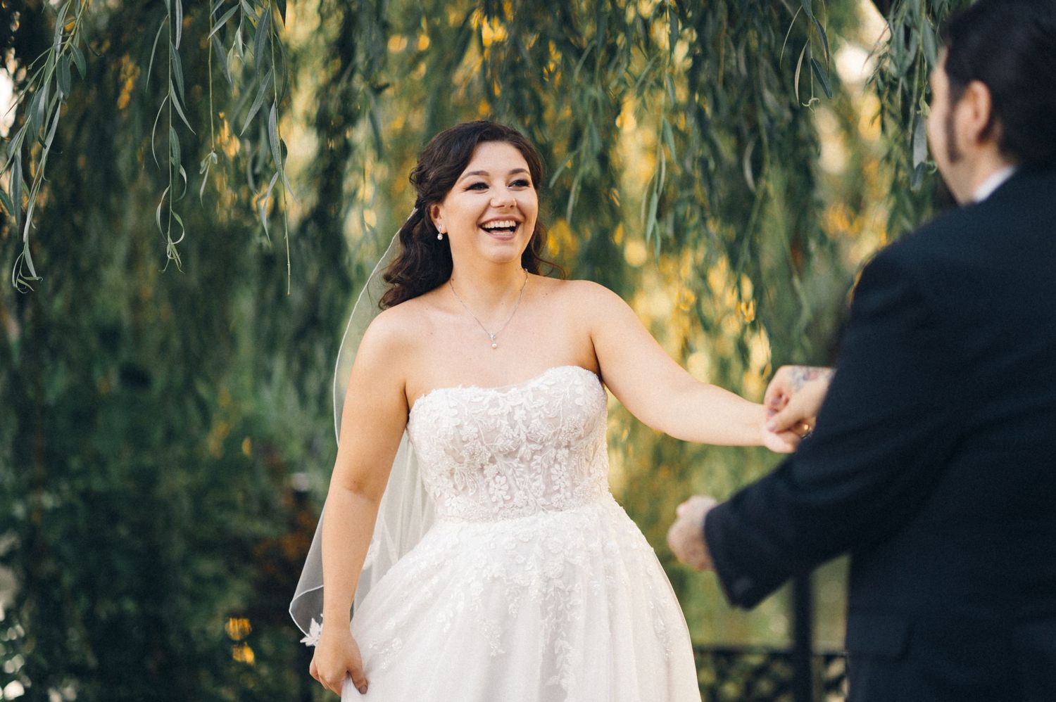 Wedding couple shares a joyful moment under weeping willow branches in natural outdoor setting.