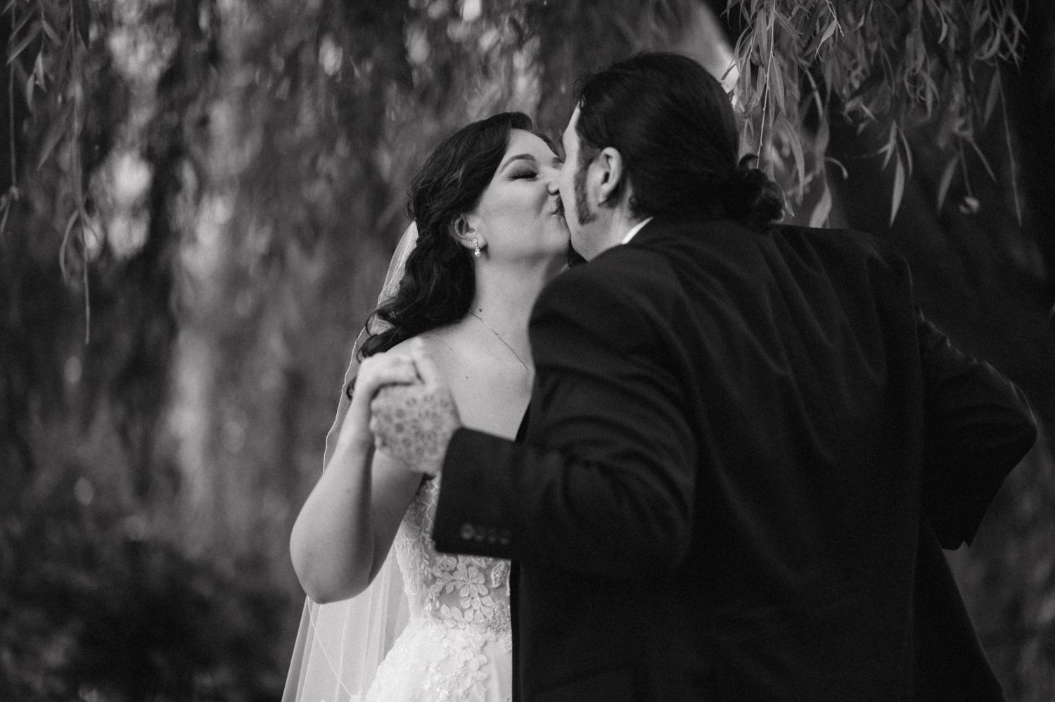 Black and white portrait of romantic wedding moment beneath weeping willow trees.