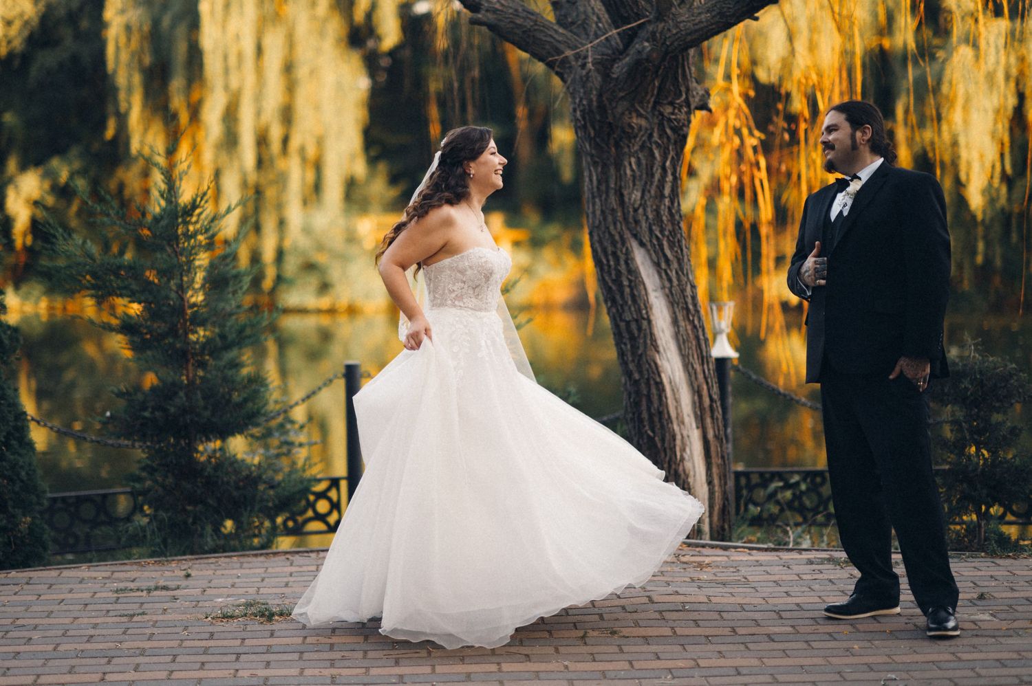 Bride in flowing white dress twirls on pathway while groom watches at golden hour.