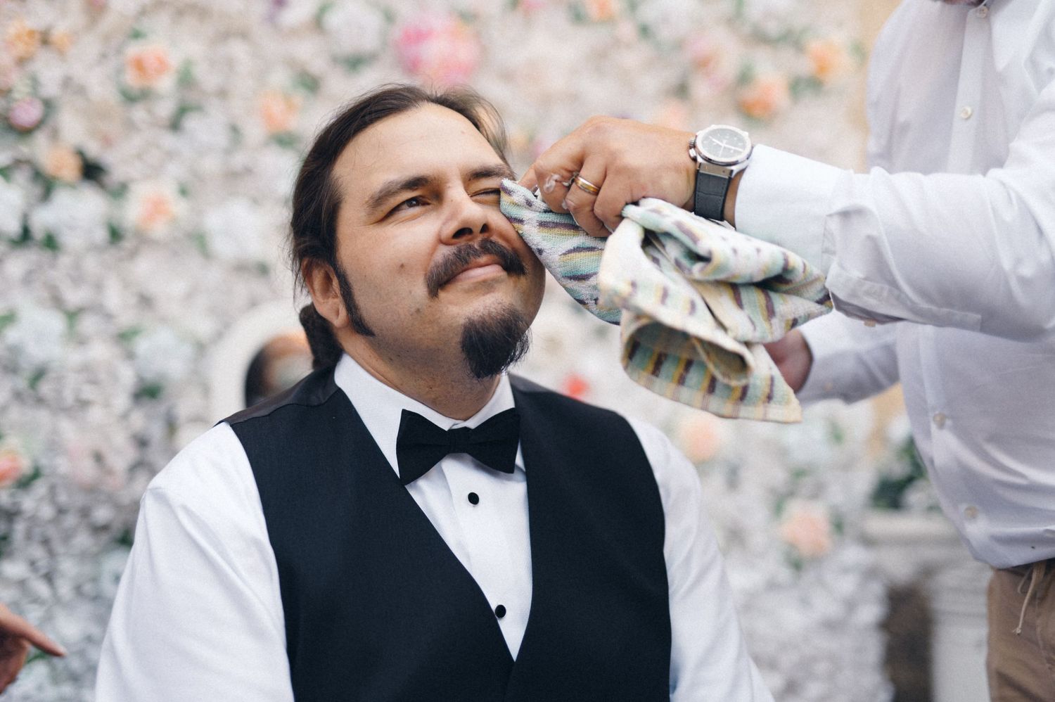 A person in formal attire getting groomed against a floral backdrop.