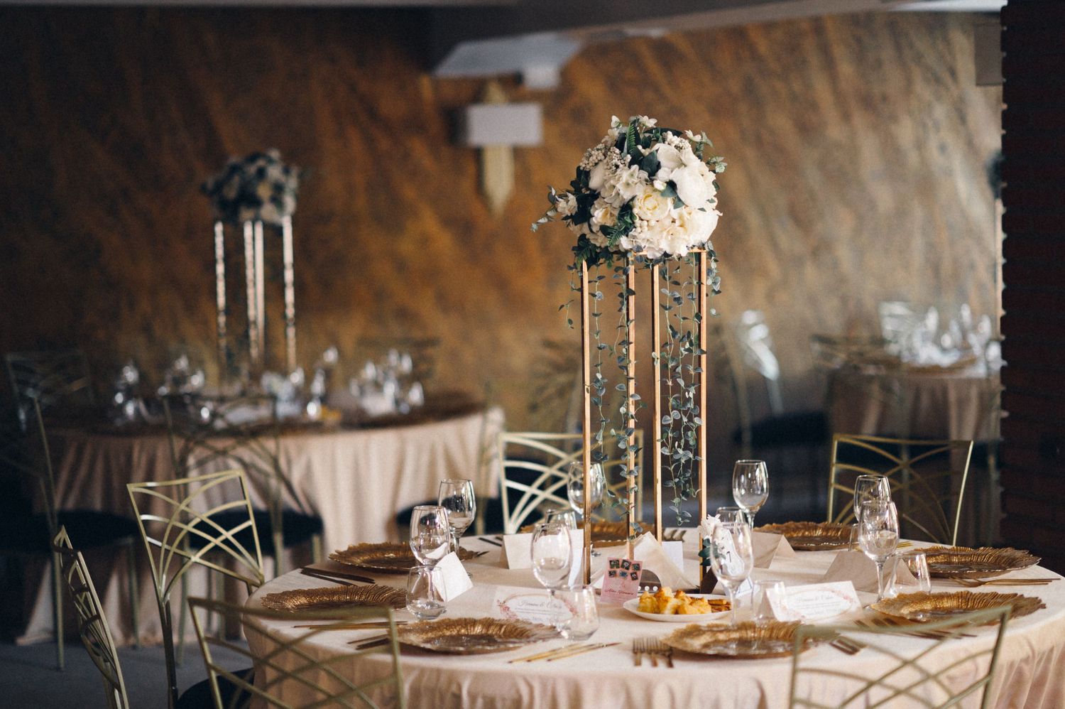 Elegant wedding reception table with tall floral centerpiece and gold accents in a rustic venue.