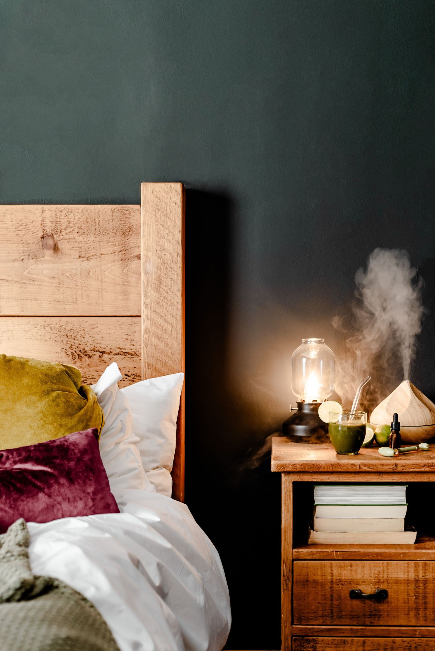 Rustic bed with red and yellow cushions against a dark green wall and a bedside lamp on with a diffuser. Cozy, inviting