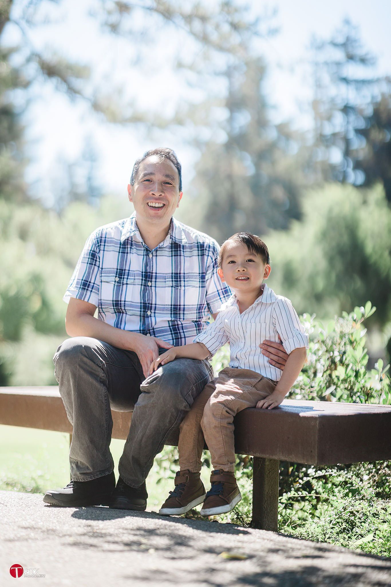 Two people sit together on a wooden bench surrounded by greenery on a bright day.